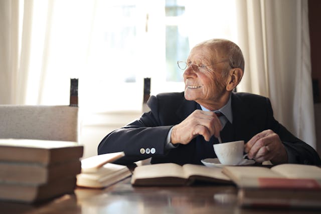 Elderly man sitting at a table, enjoying a cup of coffee while looking out the window, contemplating his surroundings.