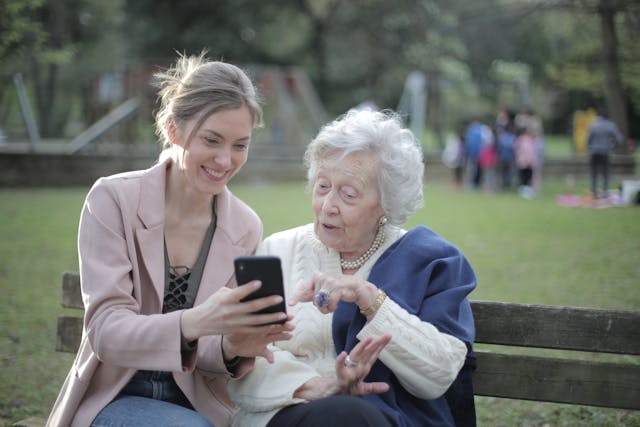 An elderly woman sitting on a bench with a younger woman, both smiling as they look at a smartphone, enjoying time together.